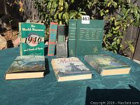 Eight vintage books arranged outdoors including 1946 World Almanac and 7 other literature novels showing spines and covers on tablecloth outdoors.