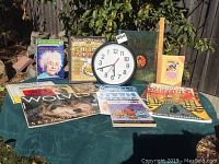 Display of books and backward clock on a green tablecloth outside showing variety of children's books and clock in front with sluggish hands.