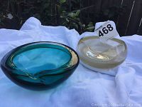 Two Murano glass ashtrays, one teal and one clear with gold speckles, displayed on white cloth outdoors.
