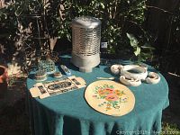 Top view of table with vintage Selectric hop heater, ceramic flower vase with mini frogs, floral art plaque, and vintage box top arranged on green tablecloth outdoors near bushes.