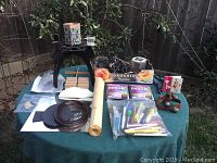 Photo of all lot items arranged on green table outdoors, showing audio equipment, lighting, crafting materials and decor items.