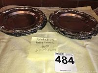 Six ornate silver plated dessert or bread plates arranged in two stacks on a pale yellow cloth with a label identifying them as Reed & Barton King Francis 1698 dessert plates.