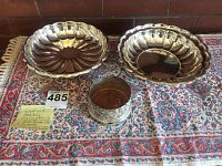 Two silver scalloped serving bowls and one silver plated carved wine bottle holder set on patterned cloth.