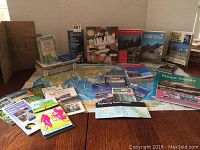 Photo showing an assortment of travel and hiking books and folded maps arranged on wooden floor, including titles about Oakland, hiking trails, Mesa Verde, California, and Point Reyes.