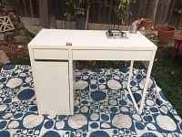 White IKEA desk shown outdoors on patterned cloth, displaying full desk with two drawers and cupboard section.