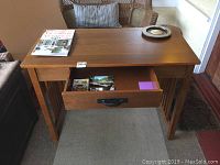 Front view of medium brown mission style wooden desk with one open drawer showing postcards and stationery on a floor mat near wicker furniture and window light.