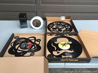 Six clocks displayed on table: three vinyl LP clocks in open boxes showing skull, gentleman, and atomic designs; two cube style black digital clocks and one small white round clock.