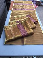 View of one curtain panel spread out showing copper and gold silk-like horizontal striped fabric with burgundy embroidered accents and mirror work, folded on outdoor table.