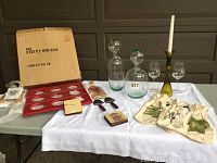 Full view of all items laid out on a white cloth including the boxed red leatherette wine rack, glass decanters, coasters, cheese plate, cheese slicers, candle holder, taper candle and cloth gift bags.