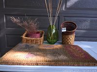 Door mat with fall leaf pattern and assorted baskets, burgundy ceramic pot, green glass vase with dried flowers on a white table with dark background
