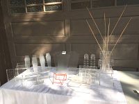 Multiple clear acrylic display pieces arranged on a white cloth including necklace stands, earring racks, risers, and an orange metal bracelet stand, photographed in daylight.
