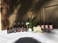 Wide view showing the full collection of 13 bath and body items and green glass vase on white tablecloth outside near garage door.