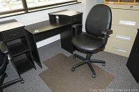 Black desk with one drawer and open shelf shown next to black vinyl office chair and chair mat on carpeted floor against windowed wall.