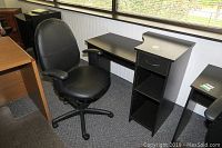 Black office chair positioned in front of a black laminate desk with side drawer and shelves. Chair mat visible beneath the chair.