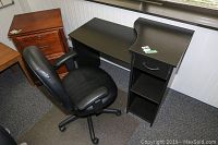 Black desk with one drawer and two shelves along with black vinyl office chair and chair mat on carpeted floor.