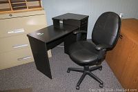Photo showing black desk with drawer and shelf next to office chair on carpet.