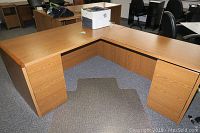 Large corner L-shaped desk with wood finish, two drawers and filing cabinet on one side, two filing drawers on the other, wood grain surface, office setting