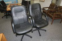 Front view of two black vinyl office chairs with armrests and swivel wheeled bases, showing seat and backrest upholstery.