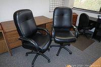 Two matching black vinyl office desk chairs seated side by side in a conference room with wooden desks in the background.