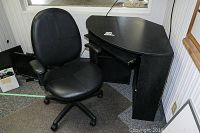 Black corner desk positioned in a corner with visible keyboard drawer slightly extended. Black vinyl office chair with armrests positioned next to the desk. Carpeted floor with visible chair mat underneath.