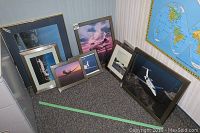 Seven framed photos of private jets arranged on floor near a corner, sizes from 10x12 to 24x20 inches. Also visible is a map on the wall behind.
