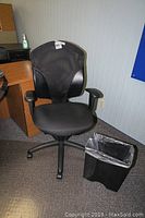 Black ergonomic office chair placed near a wooden desk with a black trash can beside it on a dark carpet.