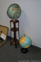 12 inch world globe mounted on a tall wooden stand and smaller globe desk lamp with black base on floor