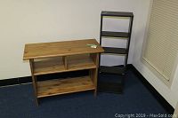 Photo showing two shelves: a natural wood bookshelf with compartments in front, and a black metal or wooden 5-tier shelf behind it, against a white wall and blue floor.