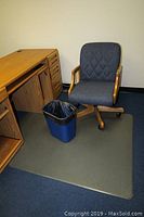 Office chair with gray fabric upholstery featuring diamond pattern, wooden arms and base, placed next to wooden desk. Blue trash can and rectangular floor mat underneath included.