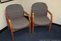Two gray fabric upholstered office accent chairs with wooden arms side by side on a carpeted floor against a wall.