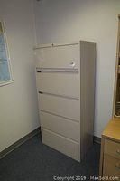 Metal lateral file cabinet with four drawers, beige color, shown from an angle in an office corner next to a desk and a bookshelf.