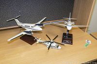 Three scale model aircraft displayed on a table, including a white Beechcraft King Air B200 airplane on a wooden stand, Falcon 50EX jet on a plaque, and a white AW139 helicopter model with black and yellow tipped rotors.