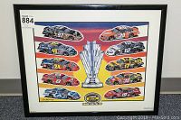 Framed colorful print showing 10 NASCAR cars surrounding a large silver trophy, set against a gradient sunset background.