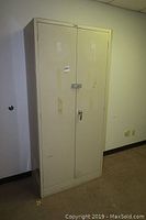 Full front view of a beige metal storage cabinet with double doors, lock and signs of wear.