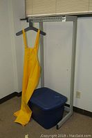 Sterilite blue plastic bin, bright yellow safety overalls hanging, and metal clothes rack in corner