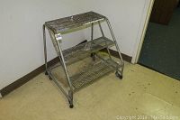 Front angled view of the metal rolling three-step ladder showing all steps, casters, and side frames.