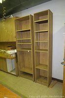 Two tall veneer wood shelves side by side, showing front view in a storage or work room environment