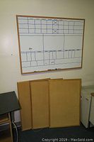 Dry erase board with grid layout mounted on the wall above three cork bulletin boards stacked on the floor.
