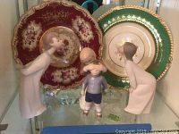 Two decorative plates on stands behind two Lladro figurines (boy and girl) on display shelf.