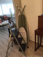 Rubbermaid three-step ladder with two ironing boards (one full size with green cover, one small with patterned cover) leaning against a wall in a room with light wood flooring.