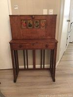 Full view of the antique wooden desk showing the drop down lid with painted artwork panels and drawer underneath.