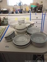 Photo showing a collection of white ceramic cooking and serving dishes on a table including two CorningWare casserole dishes with glass lids, multiple bowls, a soup tureen, and pie plates.