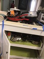Photo showing multiple skillets and pans on stove with black and red handles, and stainless steel pots stored underneath in cabinet.