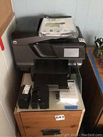 Photo of HP OfficeJet Pro 8600 Plus printer with ink cartridges and manuals on top and drawer below.