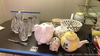 Full view of Marquis Waterford crystal pitch and cups, two piggy banks, two ceramic tea pots, and partial bowls on table.