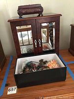Full view of the large wooden mirrored jewelry cabinet with the small black jewelry box placed in front containing assorted jewelry items.