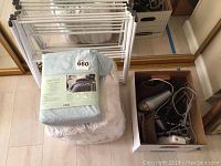 Photo of the Aircloud Vintage Wash queen sheet set package, white towels underneath, a cardboard box containing silver blow dryer, hair straightener, and two heating pads, and a white clothes drying rack in the background.