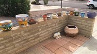 Wide view of assorted decorative pots arranged on and beside a low brick wall including large terracotta pot and small white ornamental pig decor.