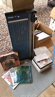 Tall blue Dahle 564 paper cutter with measuring grid, cutting arm on the right side, standing near boxes with stained glass books, paper, and craft items visible.