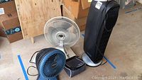 Photo of four assorted fans including a white oscillating metal blade fan, a black tower fan, a small blue personal fan, and a small black device on the floor.